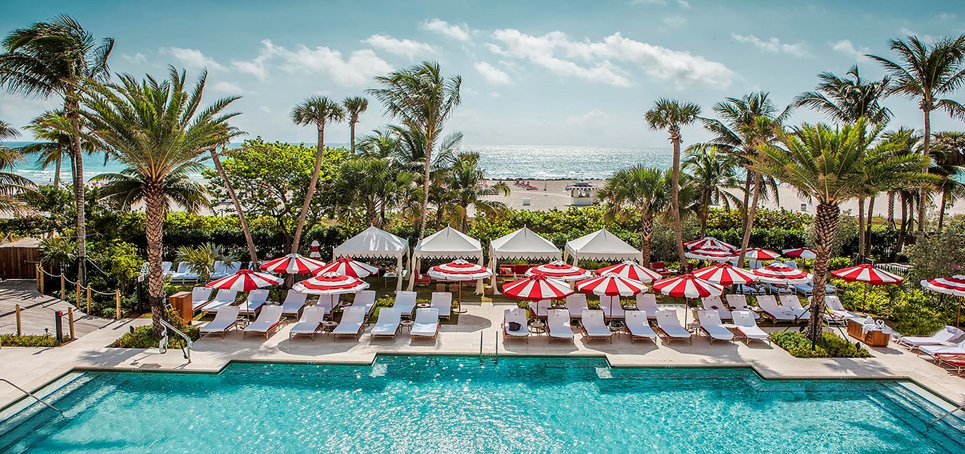 Faena Miami pool area with large pool, white lounge chairs with red and white striped umbrellas and the ocean in the background